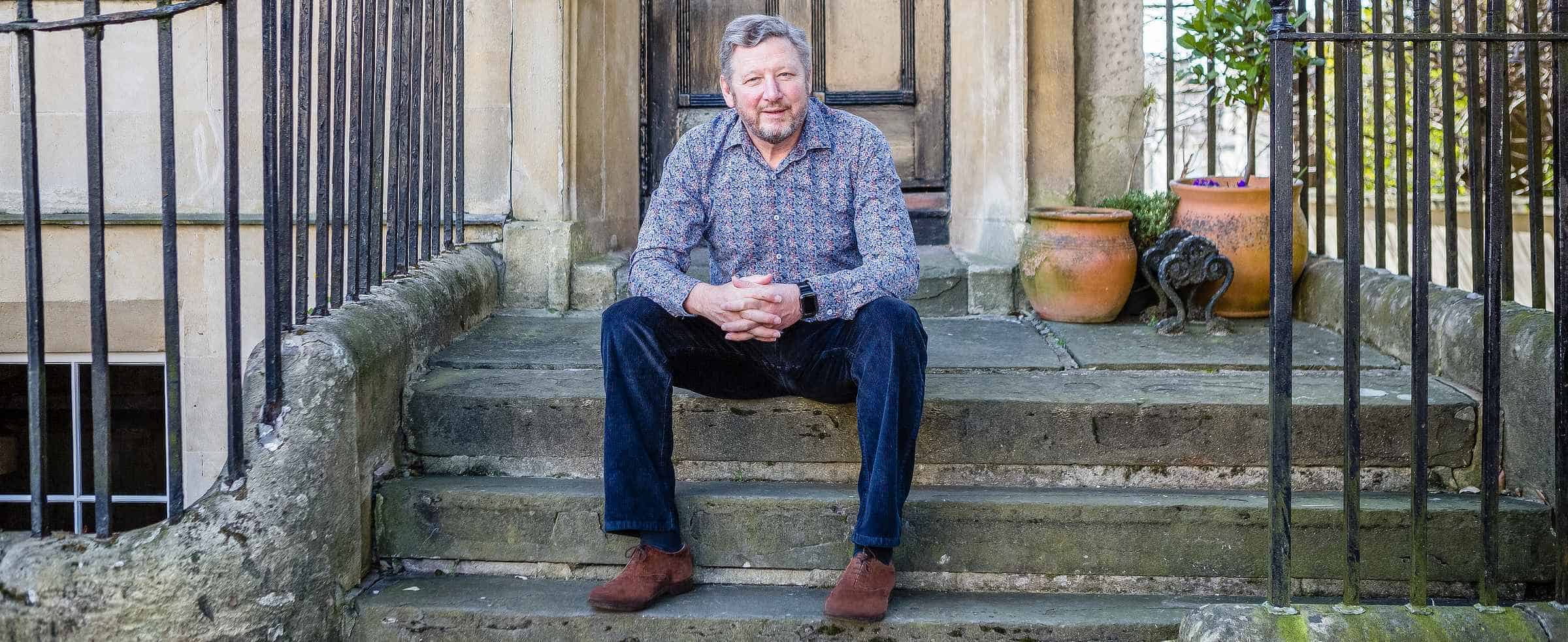 Chris Deverell, owner of Deverell Innovation Ventures, casually dressed and sitting on the front doorstep of a classic old Bath townhouse. He looks directly at the camera, hands together with fingers interlocking, exuding a relaxed yet confident demeanour.