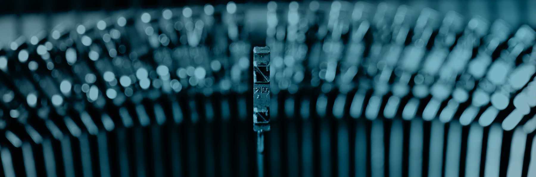 A close up, and darkened image with a navy blue filter applied, of a classic typewriter, with one type hammer in the centre of the image, coming out towards you as if it's about to print the letter on a piece of paper.